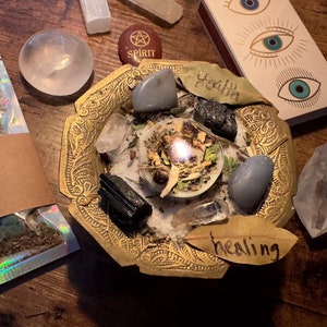 Może przedstawiać: Złota metalowa miska z różnymi kryształami, ziołami i zapaloną świecą. Wokół miski znajdują się inne kryształy, woreczek z ziołami oznaczony "Pine & Pestle House of Herbals" oraz pudełko zapałek z napisem "Health".