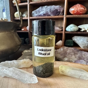 Puede incluir: Una pequeña botella de vidrio con tapa negra llena de un líquido amarillo y partículas negras. La botella tiene una etiqueta blanca que dice "Lodestone Ritual Oil".