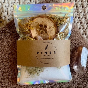 Pode incluir: Um saco de plástico transparente cheio de ervas secas, uma fatia de maçã seca e um pequeno cristal. O saco é etiquetado com "Pine & Pestle, House of Herbals".
