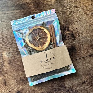 Op de afbeelding: Een doorzichtige, iriserende zak met gedroogde sinaasappelschijf, kaneelstokjes en kruiden. Een bruin papieren label met de tekst "PINE & PESTLE - HOUSE OF HERBALS" is bevestigd. De zak is afgesloten met een ritssluiting.
