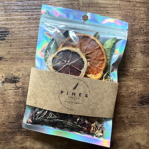 Peut inclure: Un sachet transparent et irisé rempli de tranches d'orange séchées, d'herbes et d'autres plantes. Une bande de papier brun avec le texte "PINE & PESTLE | HOUSE OF HERBALS" entoure le sachet. Le fond est une surface en bois.