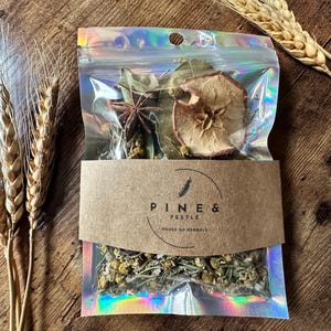 Puede incluir: Una bolsa de plástico transparente con una etiqueta de papel marrón que dice "Pine & Pestle - House of Herbals". La bolsa contiene una variedad de hierbas y especias secas, incluyendo anís estrellado, rodajas de manzana y flores de manzanilla.