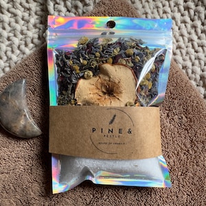 Puede incluir: Una bolsa de plástico transparente con un acabado holográfico iridiscente contiene un baño de burbujas con flores secas, hierbas y una rodaja de manzana seca. La bolsa tiene una etiqueta marrón con el texto "PINE & PESTLE - HOUSE OF HERBALS".