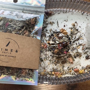 May include: A close-up of a small bag of dried herbs and flowers, labeled "Pine & Pestle - House of Herbals". The bag is open, revealing the contents, which are a mix of dried herbs and flowers. The herbs and flowers are arranged in a circle on a white surface, with a clear crystal in the center.