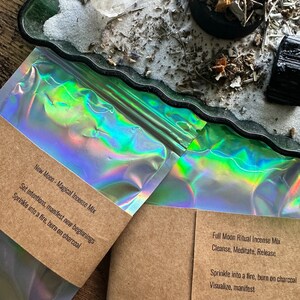 Pode incluir: Duas bolsas hologr&aacute;ficas com etiquetas de papel castanho. As etiquetas cont&ecirc;m texto que descreve o conte&uacute;do como "New Moon - Magical Incense Mix" e "Full Moon Ritual Incense Mix". O texto nas etiquetas inclui instru&ccedil;&otilde;es para usar o incenso.