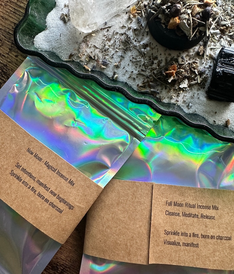 Pode incluir: Duas bolsas hologr&aacute;ficas com etiquetas de papel castanho. As etiquetas cont&ecirc;m texto que descreve o conte&uacute;do como "New Moon - Magical Incense Mix" e "Full Moon Ritual Incense Mix". O texto nas etiquetas inclui instru&ccedil;&otilde;es para usar o incenso.
