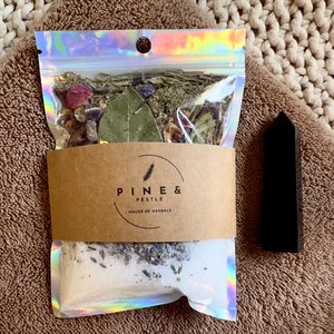 Pode incluir: Um saco de plástico transparente cheio de ervas secas, flores e cristais. O saco tem um rótulo marrom que diz "Pine & Pestle, House of Herbals". Também é possível ver uma ponta de cristal preto.