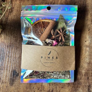 Puede incluir: Una bolsa de plástico transparente con una etiqueta de papel marrón que dice "PINE & PESTLE HOUSE OF HERBALS". La bolsa contiene una mezcla de hierbas secas, especias y frutas, incluyendo una rodaja de naranja seca, palitos de canela, anís estrellado y pétalos de rosa.