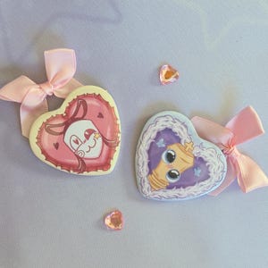 May include: Two heart-shaped badges with pink bows. One badge features a pink and red design with a cartoon face. The other has a light blue background with a cartoon chess piece. Two heart-shaped gems are scattered around.