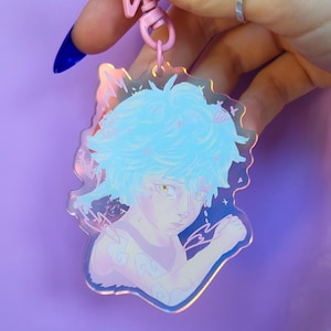 Puede incluir: Un amuleto holográfico con un personaje de pelo azul sobre un fondo rosa y blanco. El amuleto está unido a un llavero rosa y blanco.
