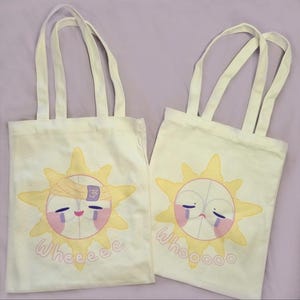 Puede incluir: Dos bolsas de mano de lona color crema con asas a juego. Cada bolsa presenta un diseño de sol de dibujos animados con una cara, lágrimas y las palabras "Wheeeee" y "Whooooo" en rosa claro. El sol tiene rayos amarillos.