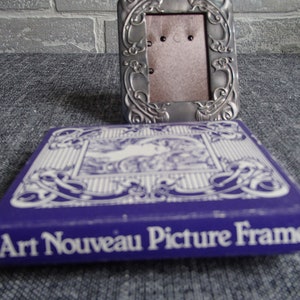 May include: A silver Art Nouveau picture frame with an ornate design. The frame is in its original packaging, which is a purple box with white lettering that reads "Art Nouveau Picture Frame".