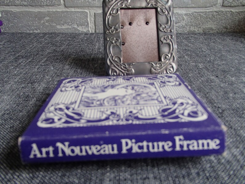 May include: A silver Art Nouveau picture frame with an ornate design. The frame is in its original packaging, which is a purple box with white lettering that reads "Art Nouveau Picture Frame".