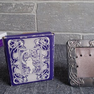 May include: A silver picture frame with an ornate design. The frame is sitting on a gray surface. The frame is empty and ready for a photo.