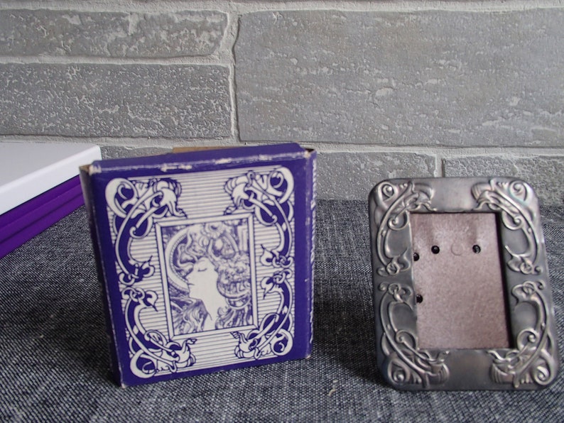 May include: A silver picture frame with an ornate design. The frame is sitting on a gray surface. The frame is empty and ready for a photo.