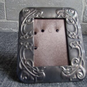 May include: A silver-toned picture frame with an ornate design featuring swirling floral motifs. The frame has a brown backing and is designed to hold a small photograph.