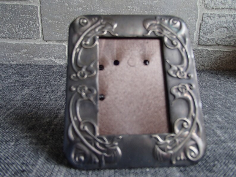 May include: A silver-toned picture frame with an ornate design featuring swirling floral motifs. The frame has a brown backing and is designed to hold a small photograph.