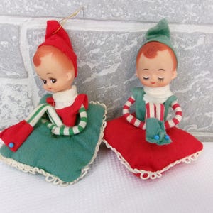 May include: Two vintage felt Christmas elf ornaments. The elves are sitting on red and green cushions. They have red and white striped sleeves and green hats. The elves have closed eyes and are smiling.