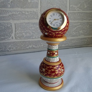 May include: A decorative clock with a round face and gold trim, set on a red and white painted base with gold accents. The base is shaped like a vase with a round top.
