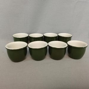 May include: Eight small, round, dark green ceramic bowls with white interiors and rims. The bowls are arranged in two rows on a neutral background. The bowls are likely for serving individual portions of food.