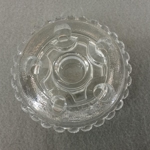 May include: A clear glass candle holder with a circular design. The holder features a central well surrounded by six smaller wells, and a scalloped edge. The glass has a textured appearance.