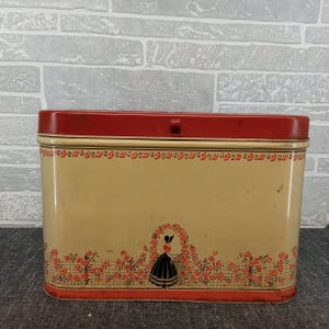 May include: A rectangular vintage tin with a red lid and base, and a cream-colored body. The body features a floral design with a woman in a black dress and a red rose border. The tin has a latch closure.