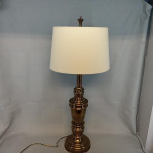 May include: A brass table lamp with a cream-colored lampshade. The lamp has a decorative base and a slender stem. The lamp is approximately 28 inches tall.