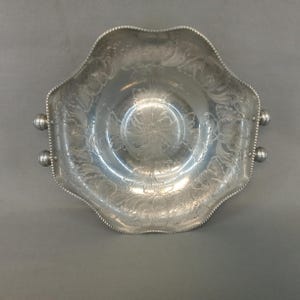 May include: A silver-toned metal serving bowl with a scalloped edge and a beaded rim. The interior surface features an etched floral pattern. Two small spherical handles are attached to opposite sides of the bowl, providing a grip for serving.