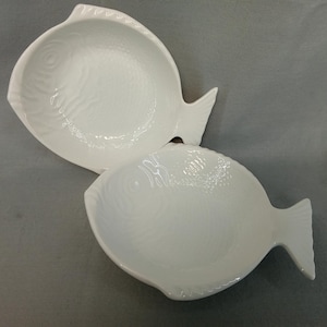 May include: Two white ceramic serving dishes shaped like fish. The dishes have a textured surface and a tail fin. The dishes are empty and are on a gray surface. The dishes are likely for serving food.