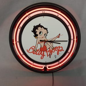 May include: A round wall clock featuring a black frame and a red neon light border. The clock face displays the cartoon character Betty Boop in a red dress. The clock hands are black, and the text "Betty Boop" is written in red.
