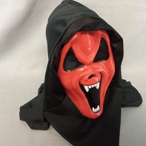 May include: A red Halloween mask with a sinister grin and white fangs, set against a black hood. The mask has black eye holes and a pointed nose. A costume accessory for Halloween or themed events.