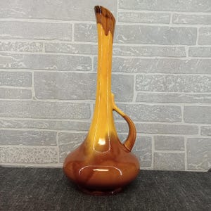May include: A tall, ceramic vase with a slender neck and a bulbous base. The vase is a gradient of colors, transitioning from a dark brown base to a golden yellow top, with a small handle. The vase is likely vintage.