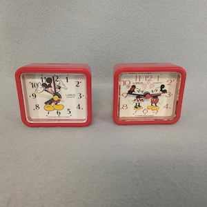 May include: Two red square Lorus quartz alarm clocks featuring Mickey and Minnie Mouse. The clocks have white faces with black numbers and hands. The Mickey Mouse clock shows Mickey pointing at the time, while the Minnie Mouse clock shows Mickey and Minnie together.
