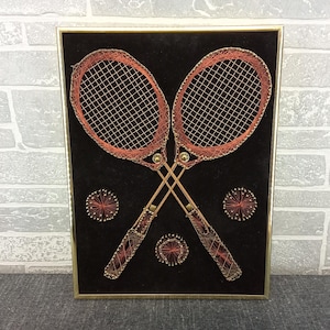 May include: Framed artwork featuring two crossed tennis rackets and three tennis balls, all crafted with red and gold thread on a black background. The rackets and balls are intricately detailed, set within a gold frame.
