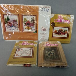 May include: Four Jiffy Stitchery and The Creative Circle embroidery kits in plastic packaging. The kits feature printed designs of a water mill, a covered bridge, a red barn, a quote about a father, and a raccoon, with text including kit numbers.