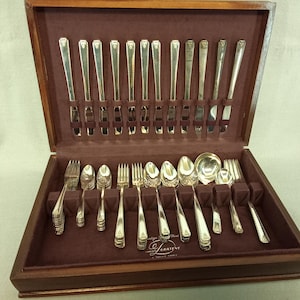May include: A wooden storage chest with a dark maroon velvet interior containing a set of silver-toned flatware. The set includes knives arranged vertically in the lid and various forks and spoons arranged horizontally in the base.