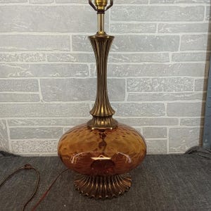 May include: A vintage table lamp with a textured amber glass shade and a brass-colored metal base and stem. The lamp has a round, bulbous shade and a fluted stem. The base is also brass-colored and has a decorative, scalloped edge.