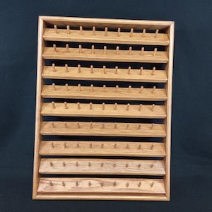 May include: A rectangular wooden wall-mounted display rack featuring eight horizontal shelves. Each shelf is equipped with a series of evenly spaced vertical wooden pegs designed for organizing and hanging small items in a grid-like arrangement.