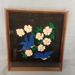 May include: Embroidered artwork in a wooden frame. The image features two blue birds perched on a branch with white flowers and green leaves against a black background. The frame is a warm brown color.