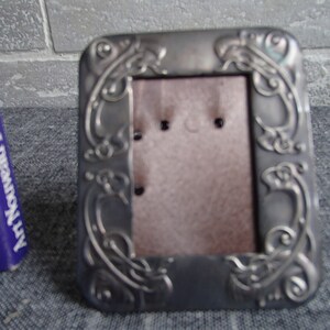 May include: A silver-toned metal picture frame with an ornate design. The frame has a rectangular shape and a stand on the back.