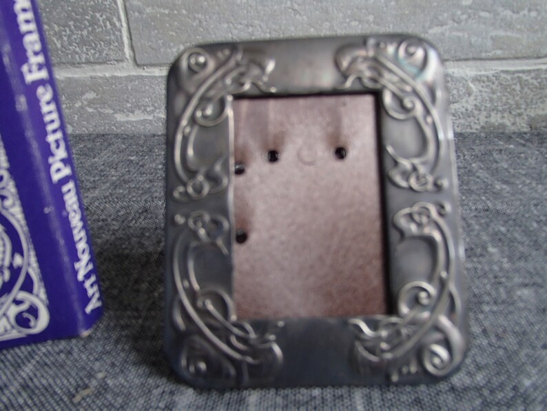 May include: A silver-toned metal picture frame with an ornate design. The frame has a rectangular shape and a stand on the back.