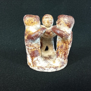 May include: A circular clay sculpture featuring three stylized figures standing in a ring, joined by their hands. The figures have a mottled, weathered finish with patches of dark reddish-brown and off-white paint over a textured surface.