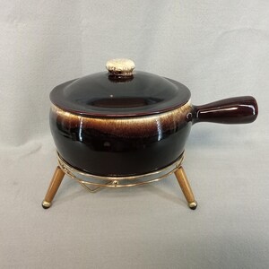 May include: A dark brown ceramic fondue pot with a matching lid and handle. The pot sits on a gold-colored metal stand with wooden legs. The lid has a small, textured knob.