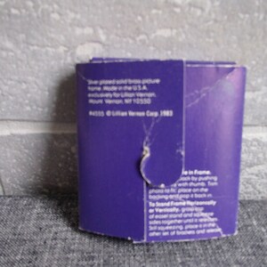 May include: Purple cardboard backing for a silver-plated solid brass picture frame. The back of the frame has instructions on how to assemble the frame and a copyright notice for Lillian Vernon Corp. 1983.
