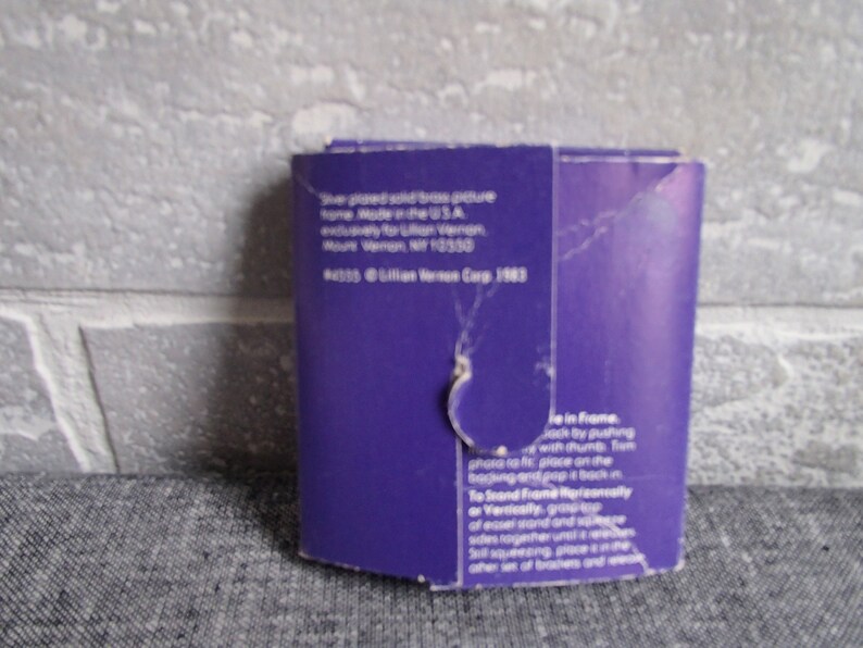 May include: Purple cardboard backing for a silver-plated solid brass picture frame. The back of the frame has instructions on how to assemble the frame and a copyright notice for Lillian Vernon Corp. 1983.