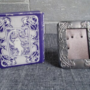 May include: A silver picture frame with an ornate design. The frame is in a box with a purple background and a white design.