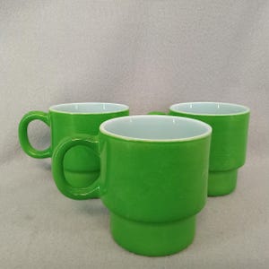 May include: Three green ceramic mugs with white interiors. Each mug has a rounded handle and a stacked base. The mugs are a bright, solid green color, and the interior is white. The mugs are designed for beverages.