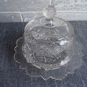 May include: A clear, cut-glass butter dish with a lid and a decorative, starburst-patterned base. The lid has a round, faceted knob. The dish sits on a scalloped, clear glass plate. The design is intricate and detailed.