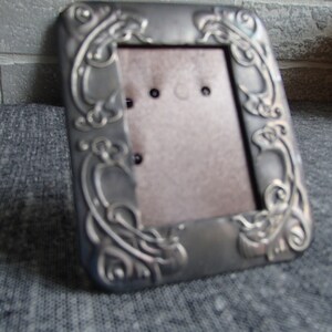 May include: A silver picture frame with an ornate design. The frame has a rectangular opening for a photo.