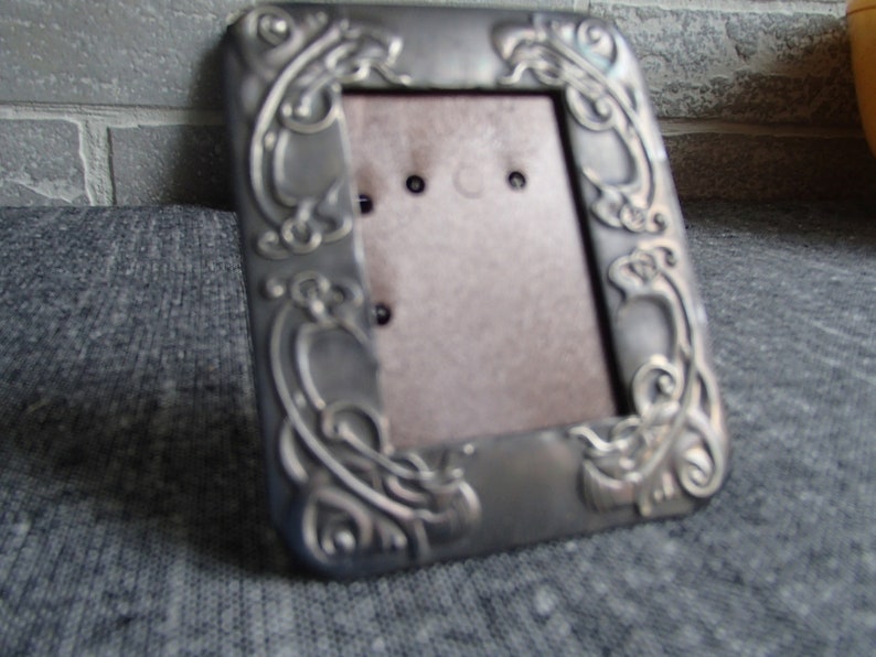 May include: A silver picture frame with an ornate design. The frame has a rectangular opening for a photo.